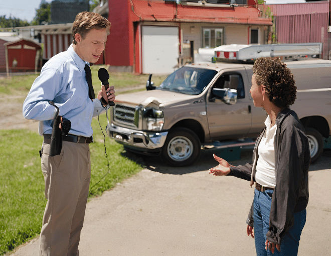A news reporter interviews a resident in front of a towing yard.